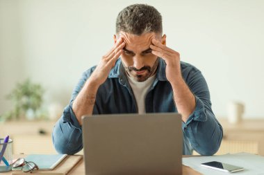 Modern ofiste dizüstü bilgisayara bakarken stres ve baş ağrısı çeken orta yaşlı bir iş adamı iş ile ilgili sorunlar yaşıyor. Çevrimiçi iş ve mesleki sorunlar