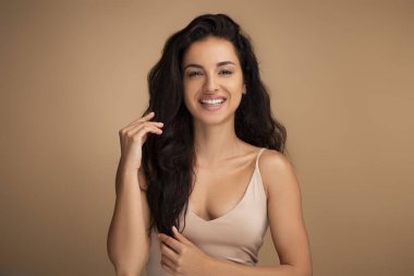 Bin yıllık mutlu esmer doğulu kadın güzel uzun saçlarına dokunuyor ve kameraya gülümsüyor, sağlıklı gür saçlarının tadını çıkarıyor, bej renkli stüdyo geçmişiyle. Saç bakımı konsepti