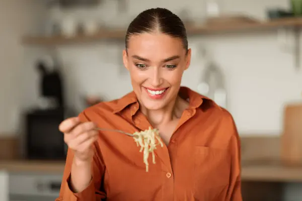 Happy woman sits in contemporary kitchen, gleefully savoring forkful of noodles, with ambient kitchen details subtly present in the background