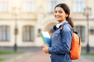 Kurs malzemeleri ve sırt çantasıyla memnun genç Hintli akademisyen bayan üniversite bahçesinde kameraya doğru ışıldıyor. Eğitim arkaplanı, boş alan.