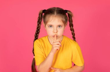 Sessiz olun. Küçük bir kızın, kameraya doğru hareketini gösteren portresi, genç bir kızın dudaklarının yanında parmağını tutması, Pembe Stüdyo Arkaplanı 'nın üzerinde dururken sessiz durması, Boşluğu kopyalaması.