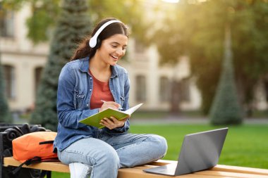 Dijital öğrenme, sanal seminer, modül. Neşeli genç Hintli bir kız öğrenci parktan online bir ders alıyor, bilgisayarını ve bluetooth kulaklıklarını kullanıyor, sanal bir test yapıyor.