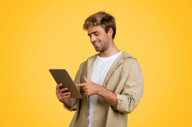 Dijital tablet kullanırken, internette sörf yaparken Avrupalı adam memnun bir şekilde ışıldıyor. Kendine güveni tam sarı bir stüdyo stüdyosunun önünde duruyor.