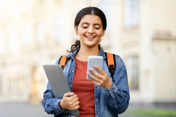 Young indian woman student texting on smartphone at campus, copy space. Positive cheerful lady with backpack and laptop in her hand walking by street, using phone, copy space. Communication