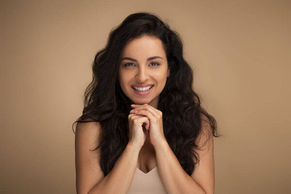 Портрет красивой брюнетки с длинными волосами тысячелетней женщины, позирующей на бежевом студийном фоне, трогающей подбородок и улыбающейся. Концепция ухода за кожей