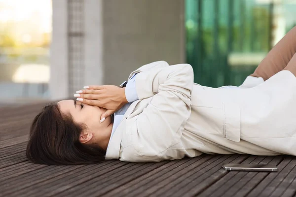 Sad tired pretty millennial lady in suit lie on floor, cry in city outdoors, near office building. Business problems, stress, bad work, overwork and depression, professional burnout