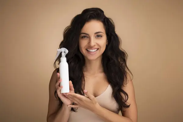 Hair care cosmetic. Cheerful beautiful millennial indian woman recommending hair care spray product on ends, split tips, treatment against dry hair or sun protection, isolated on beige background