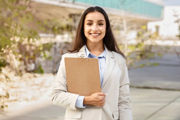 Happy pretty confident millennial woman manager in suit with clipboard enjoy work, in city office, outdoors. Business style, startup, study and lifestyle planning, exam project