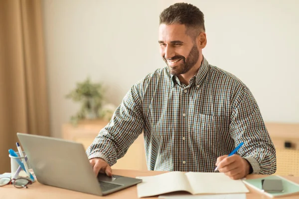 Smiling Middle Aged Man Working Online And Taking Notes Near Laptop, Sitting At Desk At Home Office Interior. Guy Websurfing Writing Business Report At Workplace. Remote Job And Entrepreneurship