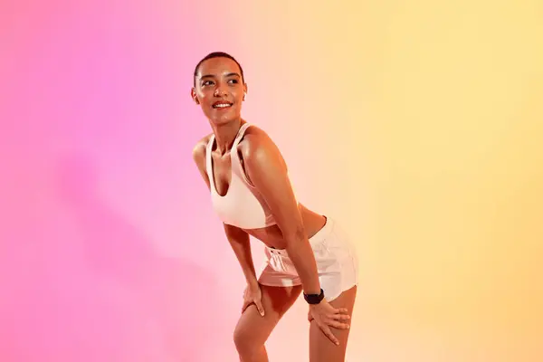 Smiling tired slim young latin lady athlete in sportswear, wireless headphones rest from run on neon pink studio background. Professional sports with music, body care, weight loss and break