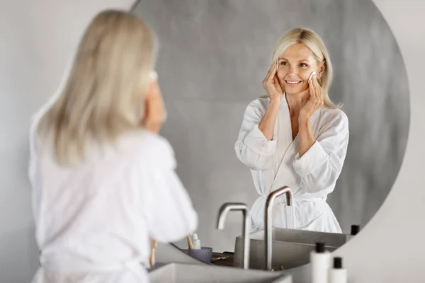 Mature Skin Routine. Smiling Senior Woman Cleansing Face With Cotton Pad And Looking In Mirror, Beautiful Elderly Lady Making Skincare Treatments In Bathroom At Home, Selective Focus On Reflection