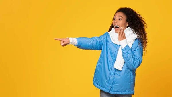 Happy and surprised black lady in winter jacket and scarf looks and points to the side at free space on yellow background, excited by great holiday offer