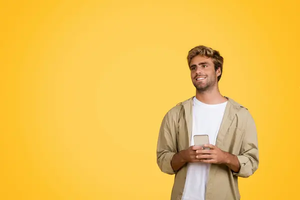 Stylish young European man holds cellphone and attentively looks towards the free space, standing against clean yellow studio background, banner