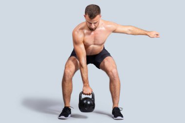 Odaklanmış, güçlü, kaslı, çıplak gövdeli beyaz bir adam spor yapıyor. Kettlebell 'e tırmanıyor. Gri stüdyo arka planında soyutlanıyor. Vücut bakımı, güç sporu, güç egzersizi.
