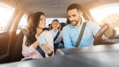 Neşeli aile araba sürmeyi sever, annesi akıllı telefon tutar, babası araba kullanır ve kızları arkada mutlu bir ifadeyle oturur.