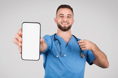 Mavi önlüklü arkadaş canlısı Avrupalı erkek doktor, beyaz ekranlı akıllı telefonu sunuyor ve onu işaret ediyor, kişiselleştirmeyi davet ediyor, gri arka plan, modelleme