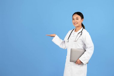 Elinde dizüstü bilgisayarla gülümseyen Asyalı kadın doktor ve avuç içi terapist bayan reklam ürünü ya da kliniği, mavi stüdyo arka planında izole edilmiş poz veren doktor kadın.