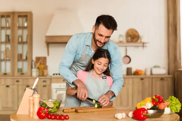 Father enjoys precious moment teaching his young daughter how to slice vegetables carefully on cutting board for healthy meal in well-equipped kitchen