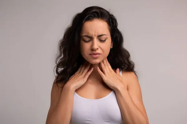 Sore throat, tonsilitis concept. Unhappy millennial brunette woman touching her neck, suffering from pain in throat, checking enlarged adenoids isolated on grey studio background, copy space