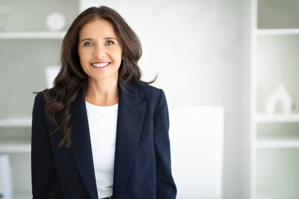 Cheerful attractive brunette middle aged woman manager posing in modern office, smiling at camera, wearing formal outfit, copy space. Career, success, business, job, occupation