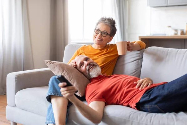 Retirement Pastime. Smiling Elderly Spouses Relaxing On Couch At Home, Happy Senior Man And Woman Watching Tv And Drinking Coffee, Enjoying Weekend Leisure, Man Lying On Wifes Laps