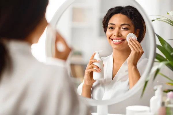 Skincare Routine. Young black woman using makeup remover and cotton pad at home, smiling african american lady sitting near mirror at dressing table, enjoying her daily skin cleanse, selective focus