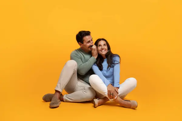 Smiling old caucasian husband whispers in ear to wife, enjoy free time, isolated on orange studio background. Family lifestyle, relationships and love, date together, secret news, gossip