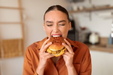 Tişörtlü kararlı kadın ağız sulandırıcı burgerden coşkulu bir ısırık alırken, ev mutfağında modern aletlerle görülebiliyor.