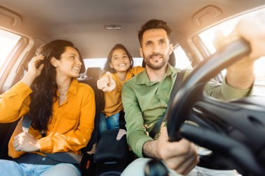 Arabanın içinde mutlu bir aile anı yakaladık. Gülümseyen baba direksiyona dokunuyor, anne saçını düzeltiyor ve kız sevinçle dışarıdaki bir şeye işaret ediyor.