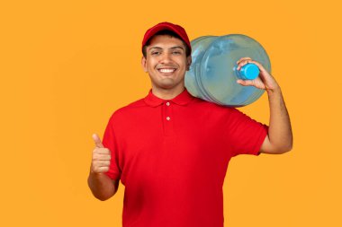 Kırmızı şapkalı ve üniformalı Orta Doğulu kurye, elinde serinlemek için temiz su şişesi tutuyor. Stüdyoda sarı arka planda poz veriyor. Onaylama jesti ile internet üzerinden bedava teslimatı onaylıyor.