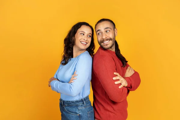 Positive millennial man and woman with arms crossed on chest standing back to back, looking at each other and smiling, isolated on yellow studio background, couple friends posing together