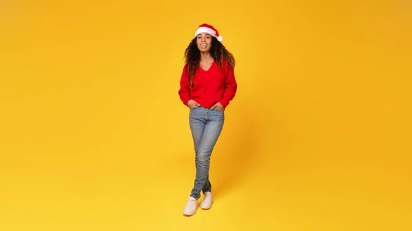 Happy young black lady wearing Santa hat and posing with hands in pockets, full-length shot against yellow background, panorama, free space