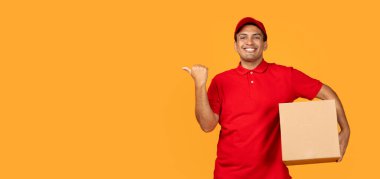 Kırmızı şapkalı ve üniformalı Latin kurye, karton paket kutusu tutuyor, dağıtım servisi reklamı için boş yeri işaret ediyor, sarı stüdyo arka planının üzerinde duruyor. Bedava reklamı olan Panorama