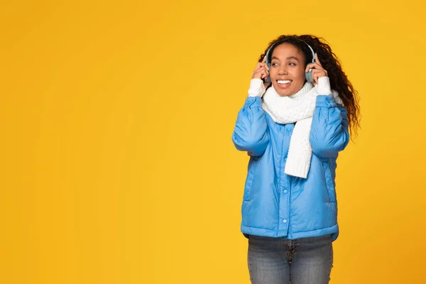 Happy young black lady in winter jacket and scarf touches her headphones and looks aside at free space, enjoying music on yellow background, holiday vibe