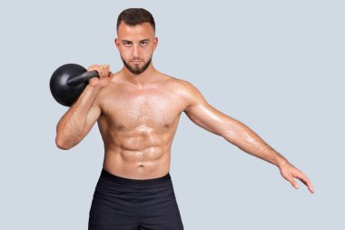 Yoğunlaşmış ter güçlü kaslı beyaz adam sakallı, çıplak gövdeli, kettlebell ile egzersiz yapan, güç antrenmanı yapan, gri stüdyo arka planında izole edilmiş. Vücut bakımı, güç sporu