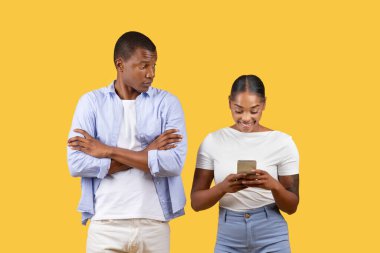 Meraklı Afrikalı Amerikalı bir adam, akıllı telefonuna dalmış, parlak sarı arka fona karşı birlikte duran gülümseyen bir kadına bakıyor..