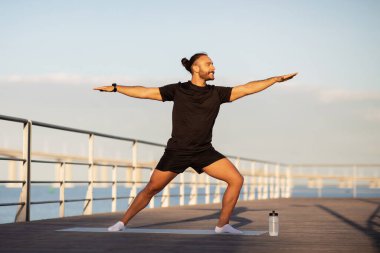 Sportif ve dengeli yoga yapan genç bir adam kollarını açmış, deniz iskelesinde savaşçı pozunda duruyor, sabah deniz kenarında spor ve doğanın ahengi ile eğleniyor.