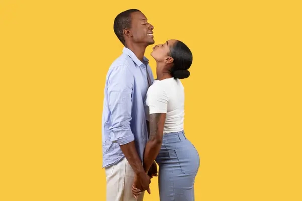Black couple standing close, woman looking up lovingly at her man and trying to kiss him as they hold hands against warm yellow backdrop, radiating intimacy and affection