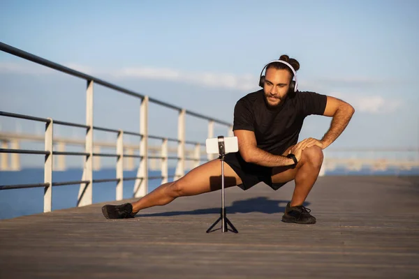 スポーツブログ 深いサイドラウンジのミレニアルフィットネス男は 三脚でスマートフォンを介して運動をストリーミングし 海辺で屋外でトレーニングします 技術と健康的なアクティブエクササイズ — ストック写真