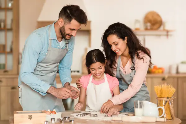 Joyful european family of three engaged in baking cookies, with young girl pressing cookie cutters into dough as her parents lovingly assist
