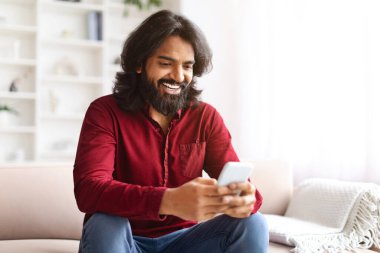 Mutlu, uzun sakallı, genç Hintli adam evinde dinleniyor, rahat bir oturma odasında koltukta oturuyor, yepyeni bir cep telefonu kullanıyor, kızlarla flört uygulamasında sohbet ediyor, kaydırıyor, kopyalama yapıyor.