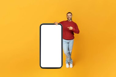 Online teklif konsepti. Neşeli, uzun saçlı, gündelik bir şekilde sarı stüdyo arka planında beyaz beyaz ekranlı akıllı telefonu işaret eden bin yıllık adam.
