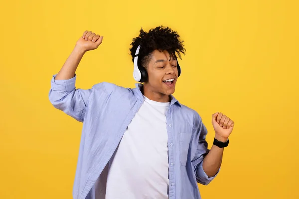 Joyful black guy with curly hair feeling the rhythm, dancing with closed eyes and headphones, expressing success and happiness on sunny yellow backdrop