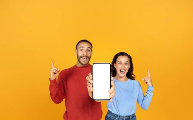 Güzel, şaşkın, mutlu bir milenyum çifti ellerinde beyaz ekranları ve sarı stüdyo arkaplanı olan akıllı telefonlarıyla, maketlerini yapıyorlar. İki kişilik güzel bir anlaşma, harika bir online teklif.
