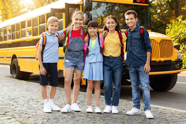 Cheerful kids with backpacks standing by yellow school bus, smiling after day of learning, group of joyful children embracing and looking at camera, ready to head home, full length shot