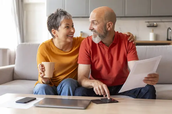 Happy Senior Spouses Checking Financial Documents And Calculating Family Budget At Home, Cheerful Elderly Couple Sitting At Table In Living Room, Reading Loan Papers Or Counting Monthly Expenses
