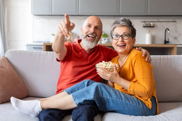 Cheerful senior spouses watching tv and eating popcorn at home, happy elderly couple having fun while relaxing on couch in living room, laughing together and pointing at camera, closeup