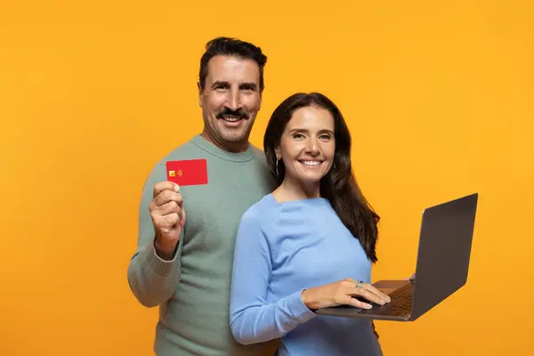 Cheerful senior european family use laptop, show credit card, isolated on orange studio background. Recommendation sale, money finance, pay for order online, shopping, cashback