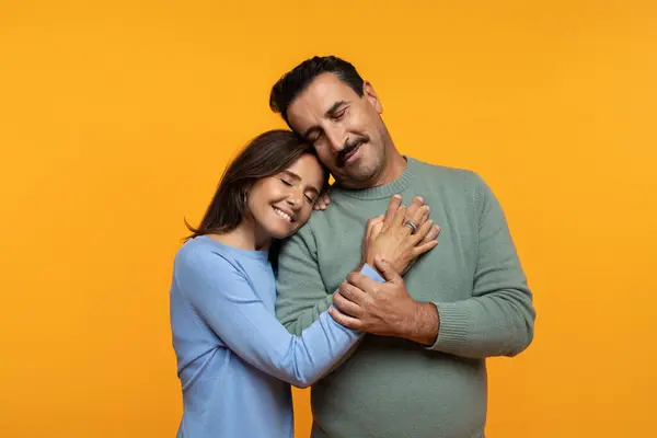 Smiling calm senior caucasian man and woman hug, with closed eyes, enjoy spare time together, isolated on orange studio background. Family lifestyle, relationships and love emotions