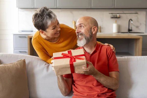 Pleasant Surprise. Loving senior woman giving gift to her husband sitting on couch at home, smiling older lady surprising her happy spouse while they resting together in living room , copy space